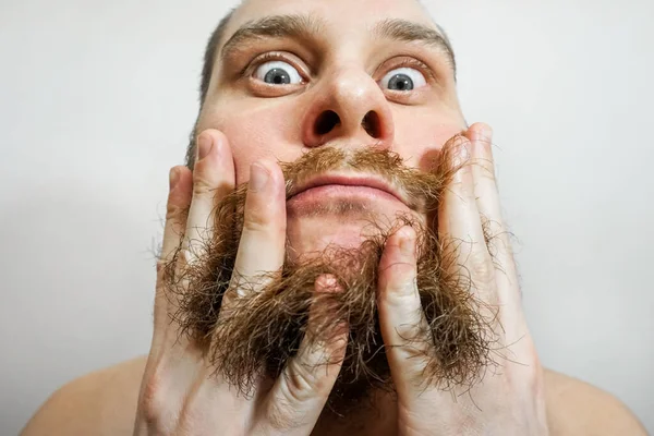 Nahaufnahme eines jungen Mannes, der sich seinen Bart mit einem Kamm frisiert, während er allein in einem Studio vor grauem Hintergrund steht — Stockfoto