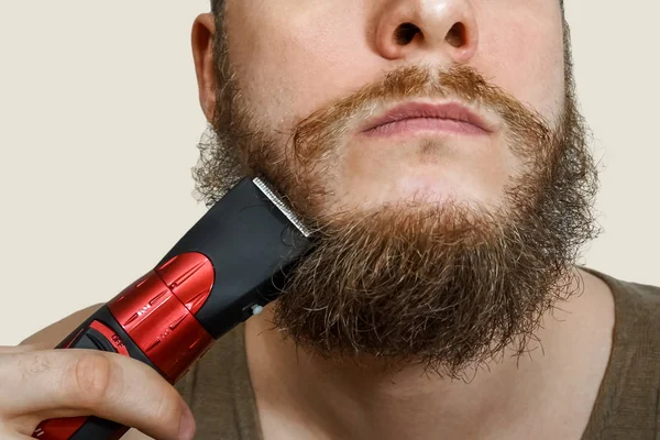 Portrait of a bearded guy cuts his beard, hair on his head and mustache with a clipper on an isolated background — Stock Photo, Image