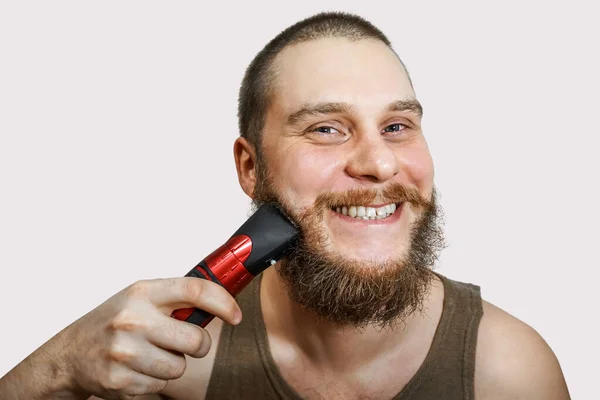 Portré egy szakállas fickóról, aki levágja a szakállát, a haját és a bajuszát egy ollóval egy elszigetelt háttérben. — Stock Fotó