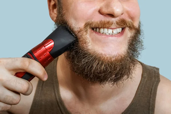 Portrait of a bearded guy cuts his beard, hair on his head and mustache with a clipper on an isolated background — Stock Photo, Image