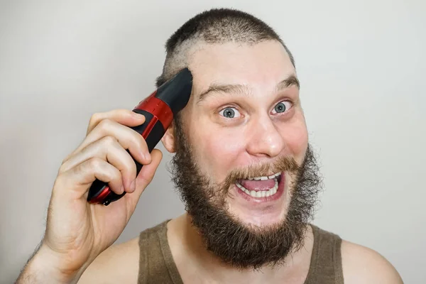 Portrait of a bearded guy cuts his beard, hair on his head and mustache with a clipper on an isolated background — Stock Photo, Image