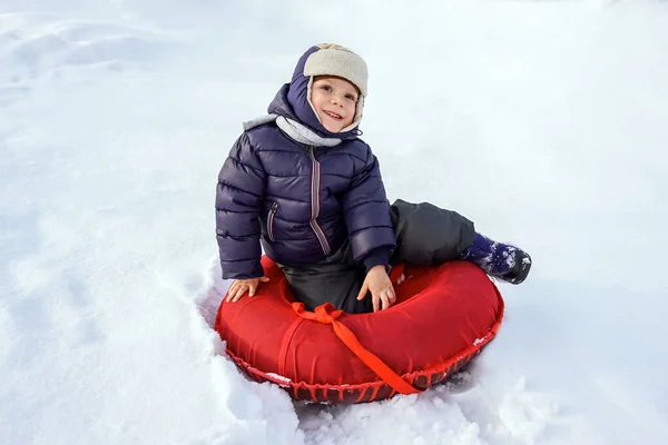 Happy joyful beautiful child boy rides from the mountain on a red tubing in winter — Stock Photo, Image