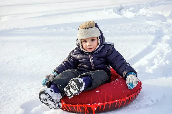 Happy joyful beautiful child boy rides from the mountain on red tubing in winter — Stock Photo, Image