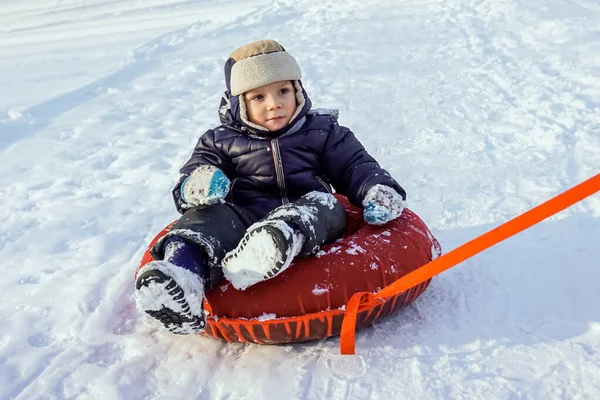 Happy joyful beautiful child boy rides from the mountain on red tubing in winter — Stock Photo, Image
