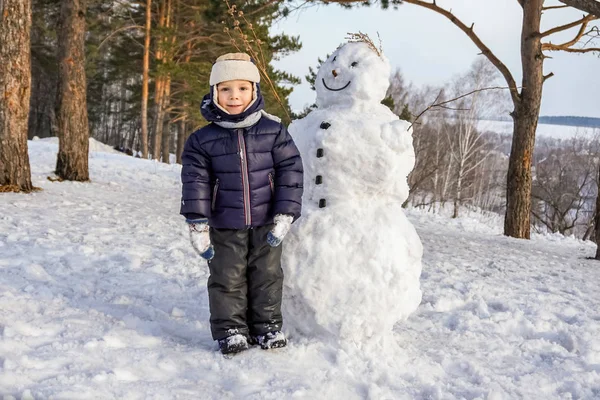 Kleiner Junge im Winterspaß mit Schneemann. Aktive Freizeit im Freien mit Kindern im Winter. Kind mit warmer Mütze, Handschuhen — Stockfoto