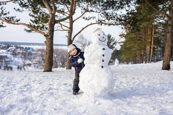 Little boy in winter fun with snowman. Active outdoors leisure with children in winter. Kid with warm hat, hand gloves