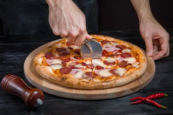 Cortar pizza picante con salami, carne, jamón, salchicha, pimientos, tomate, queso con cortador en una tabla de madera — Foto de Stock