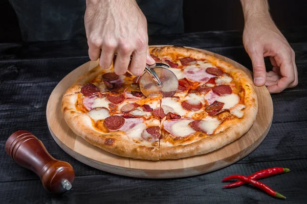 Cortar pizza picante con salami, carne, jamón, salchicha, pimientos, tomate, queso con cortador en una tabla de madera — Foto de Stock