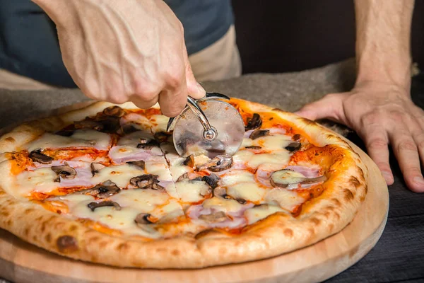 Cortar pizza picante con salami, carne, jamón, salchicha, pimientos, champiñones, tomate, queso con cortador en una tabla de madera — Foto de Stock