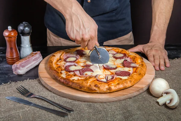 Cutting spicy pizza with salami, meat, ham, sausage, peppers, mushrooms, tomato, cheese with cutter on a wooden board — Stock Photo, Image