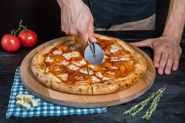 Cortar pizza picante con salami, carne, jamón, salchicha, pimientos, champiñones, tomate, queso con cortador en una tabla de madera — Foto de Stock