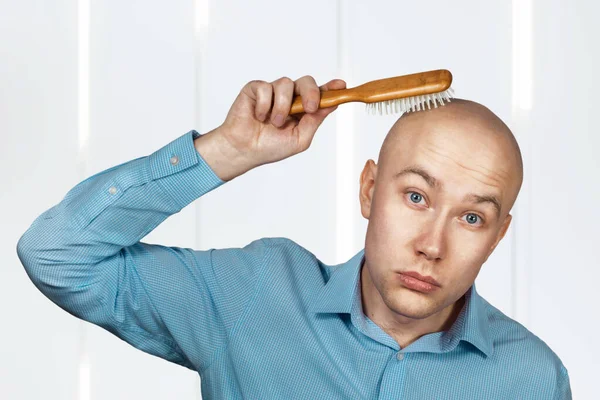 Portrait of a bald guy in a blue shirt holding a comb in his hands. The concept of hair loss and hair transplantation.