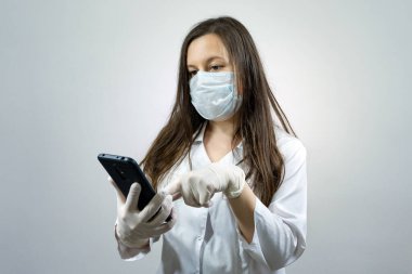 Doktor kadın koruyucu maske takıyor ve içeride eldiven giyiyor. Cep telefonu kullanıyor. Corona virüsü konsepti.