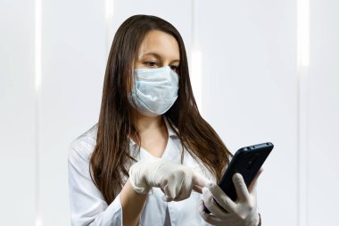 Doktor kadın koruyucu maske takıyor ve içeride eldiven giyiyor. Cep telefonu kullanıyor. Corona virüsü konsepti.