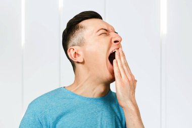 Genç, uykulu, esneyen adam elini uzatmış, mavi tişört giymiş..