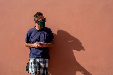 Tıbbi maskeli olgun bir adam elinde cep telefonu tutuyor.,