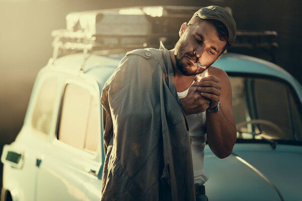 Mature bearded man lighting a cigarette, a retro car with suitcases on a roof rack on background