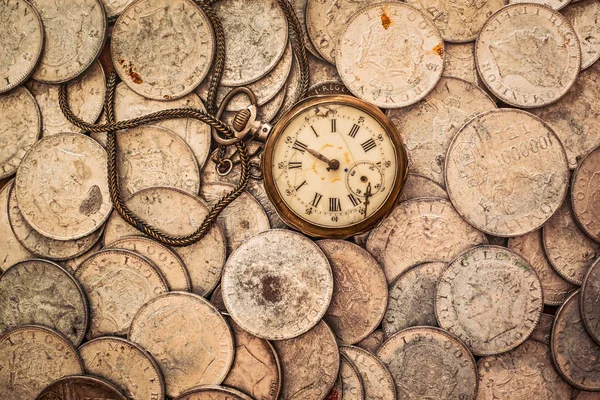 A bunch of coins with a broken pocket watch on top, top view