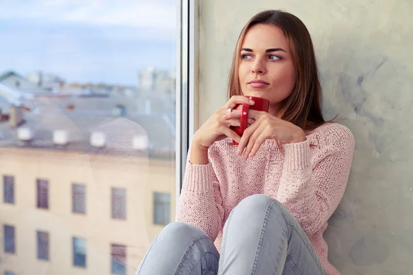 Myšlení mladá žena na parapetu s šálkem čaje — Stock fotografie