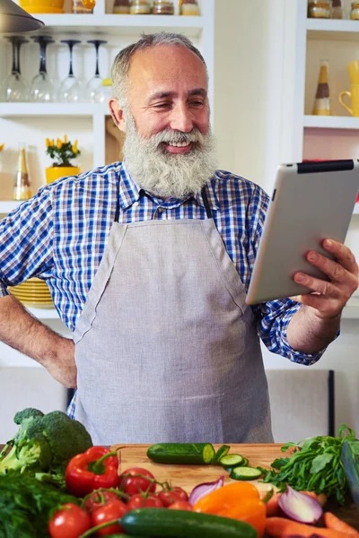 Cheerful man wearing gray apron using tablet to find a recipe