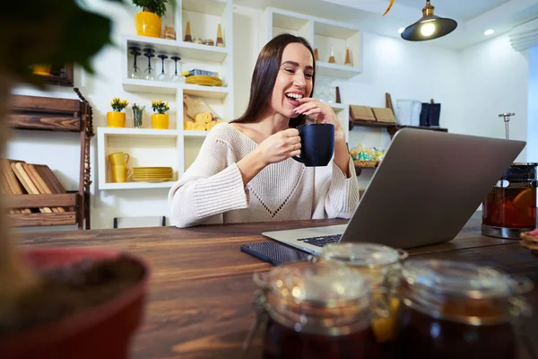 Riendo joven mujer que trabaja en el ordenador portátil mientras sostiene una taza de te —  Fotos de Stock