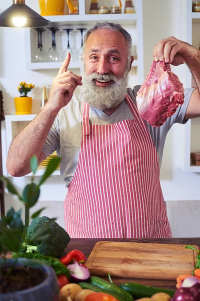 Homem com um pedaço de carne crua a gesticular o polegar na cozinha — Fotografia de Stock