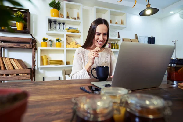 Zufriedene Frau hält einen Keks in der Hand, während sie einen Laptop benutzt — Stockfoto