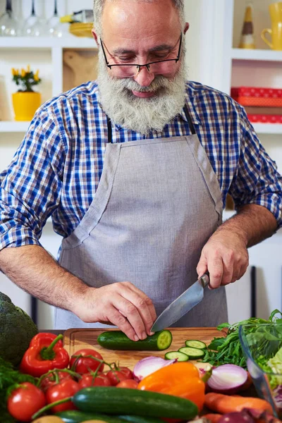 Portrait d'un homme âgé portant un tablier gris coupant le concombre tout en — Photo
