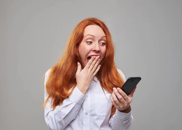 Red hair teen looking at screen of mobile phone and covers mouth — Stock Photo, Image