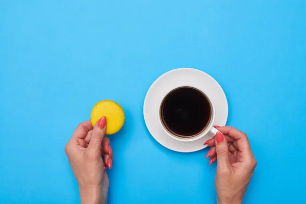 Manos sosteniendo un macarrón y una taza de café sobre una plancha azul — Foto de Stock