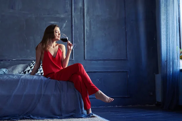 Smal vacker kvinna med ett glas vin sitter på sängen — Stockfoto