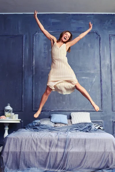 Extremely excited woman in nightgown jumping on bed with arms an — Stock Photo, Image