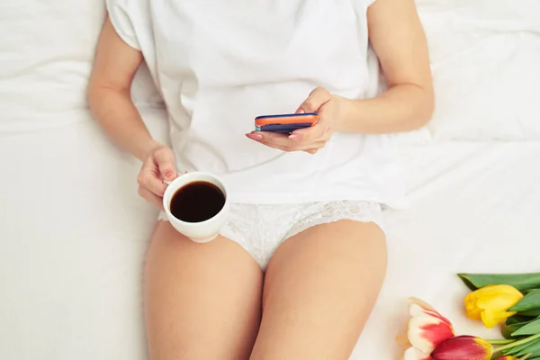 Menina com uma xícara de café usando telefone enquanto deitado em um branco — Fotografia de Stock