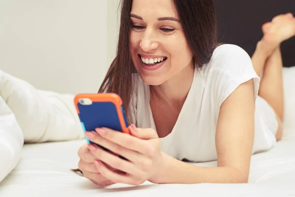 Glad kvinna skriver på mobilen medan du ligger på sängen — Stockfoto