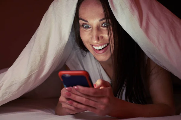 Mujer asombrada escondida debajo de la manta por la noche con un pho móvil —  Fotos de Stock