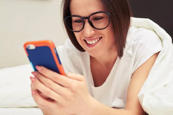 Smiling brunette in glasses relaxing on the bed and using her ph — Stock Photo, Image