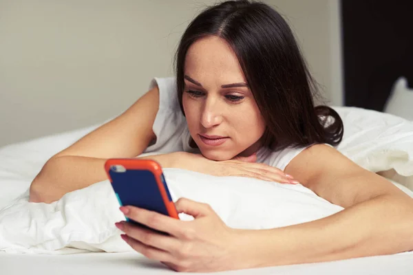 Eftertänksam kvinna ser ut på skärmen i telefon medan ligg — Stockfoto