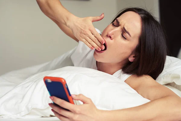 Morena bostezando en la cama y sosteniendo un teléfono móvil —  Fotos de Stock