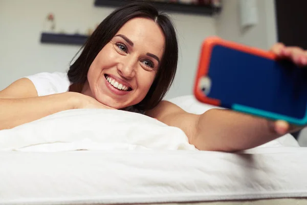 Toothy sorrindo fêmea fazendo selfie enquanto deitado na cama — Fotografia de Stock