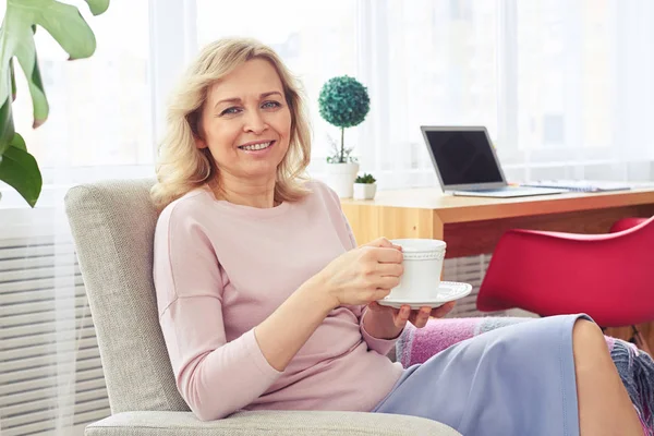 Atractiva mujer sonriendo y bebiendo café mientras se sienta en tan —  Fotos de Stock