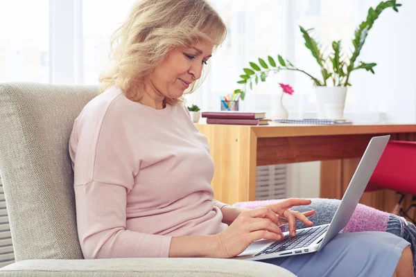 Ganska blond kvinnlig utskrift i laptop medan du sitter i fåtölj — Stockfoto