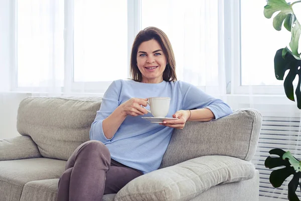Улыбающаяся мадам, отдыхающая на диване с чашкой кофе — стоковое фото