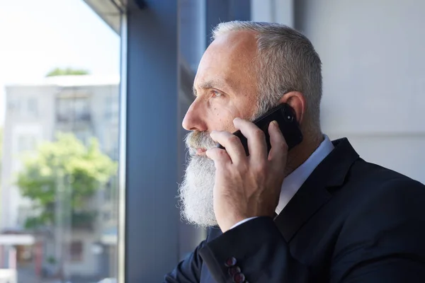 Velho senhor com barba cinza falando ao telefone enquanto olha para fora windo — Fotografia de Stock