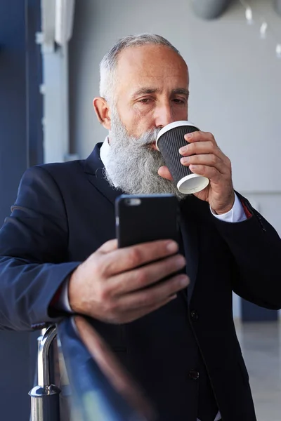 Barbudo idade masculina de 50-60 beber café e surf no telefone — Fotografia de Stock