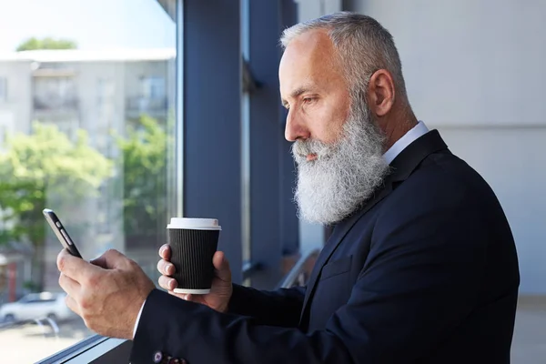 Gris señor edad de 50-60 taza de café de celebración y surf en el teléfono —  Fotos de Stock