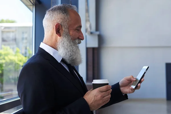 Sonriente anciano bebiendo café y surfeando por teléfono —  Fotos de Stock