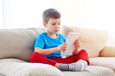 Evdeki kanepede tablet kullanan küçük çocuk.