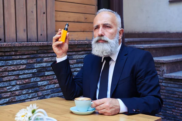 Señor con barba gris bebiendo café mientras fuma electrocigarro —  Fotos de Stock