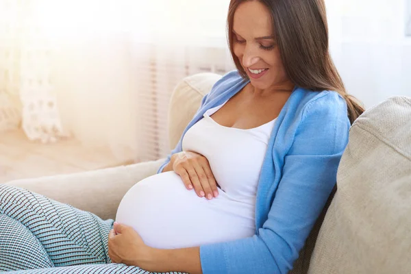 Kobieta w ciąży, trzymając jej brzuch z ramionami na kanapie — Zdjęcie stockowe