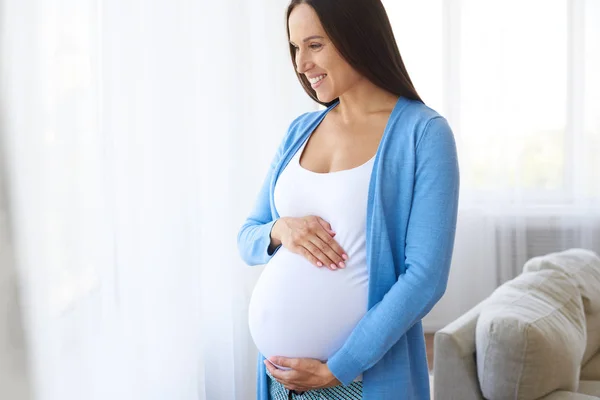 Kobieta w ciąży dotykając brzucha w pobliżu okna — Zdjęcie stockowe